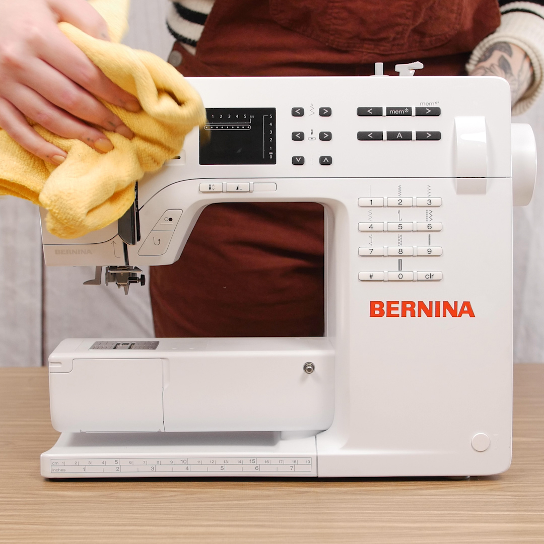 A hand using a dry cloth to wipe dust from the exterior surfaces of a sewing machine, including the arm and body