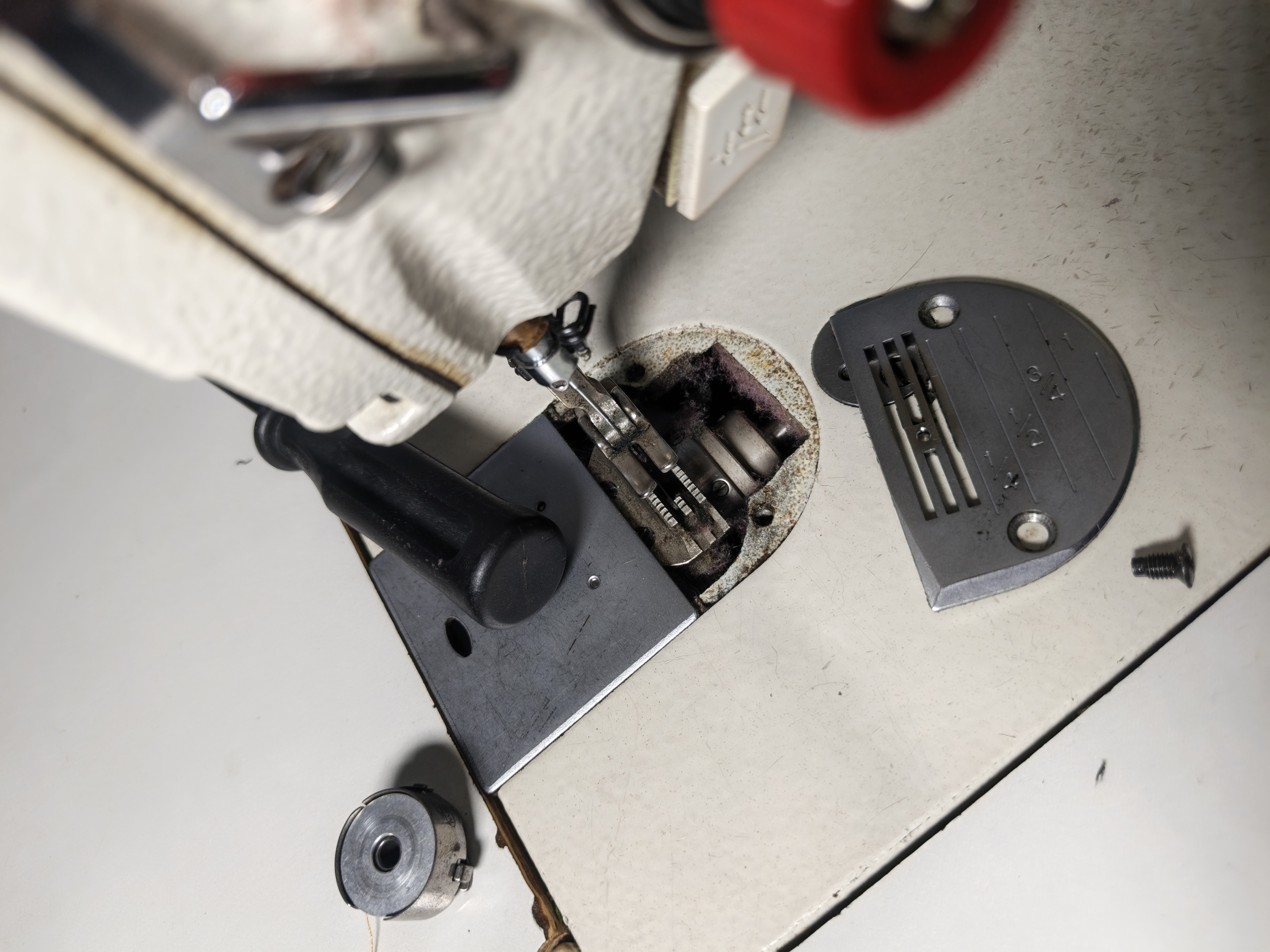 Close-up of a hand using a screwdriver to remove the needle plate, revealing lint-covered feed dogs beneath