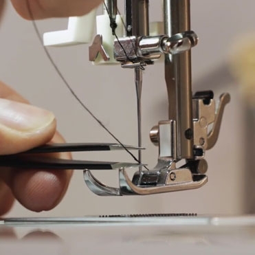 Close-up of a domestic sewing machine with presser foot raised, showing a contrasting thread tracing the correct path from the spool pin through each guide, the tension discs, and the take-up lever